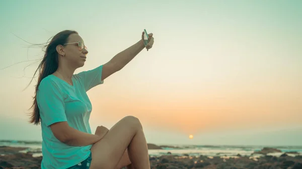 Uma bela jovem está tomando selfie em um telefone celular, sentado nas rochas junto ao mar contra o pôr do sol . — Fotografia de Stock