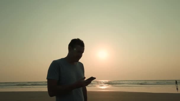 Handsome homem de pé e falando em smartphone na costa. Retrato de homem agradável passar tempo desfrutando de férias e falar em telefone celular na praia contra o pôr do sol. — Vídeo de Stock