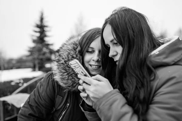 A reunião de namoradas na rua. Duas mulheres olham no celular, considerando suas fotos a pé. Foto em preto e branco. — Fotografia de Stock