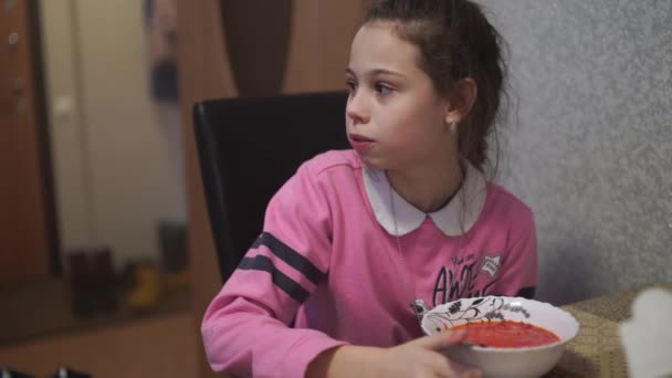 Le petit enfant mange du borscht. Charming girl chews food, talks and laughs, sitting at table in kitchen. — Video