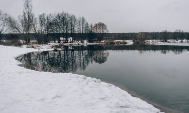 Soğuk kış gününde sakin bir gölet. Huzurlu göl suyu ve karlı sahil soğuk gri bir günde kırsal kesimde