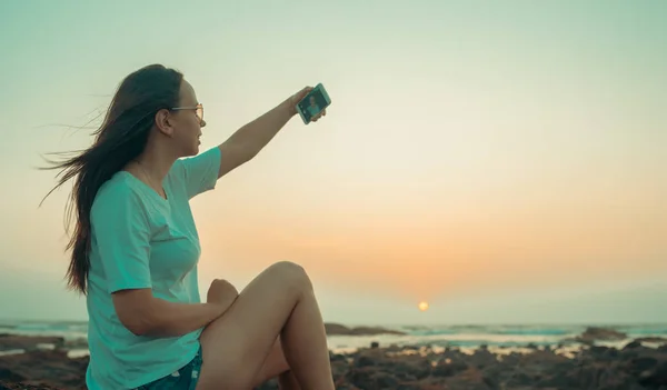 Μια όμορφη νεαρή γυναίκα παίρνει selfie σε ένα κινητό τηλέφωνο, κάθεται στα βράχια δίπλα στη θάλασσα με θέα το ηλιοβασίλεμα. — Φωτογραφία Αρχείου