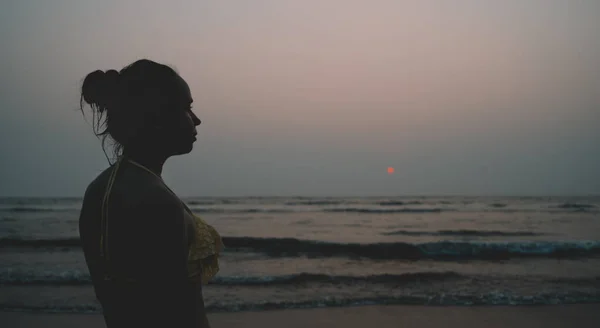 Femme anonyme près de la mer au coucher du soleil. Back view of unrecognizable female in swimwear standing on beach near waving sea in evening on resort — Photo