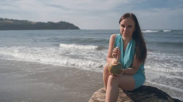 Una hermosa joven está sentada en un tronco y bebiendo el coco junto al mar o al océano en un día soleado . — Foto de Stock