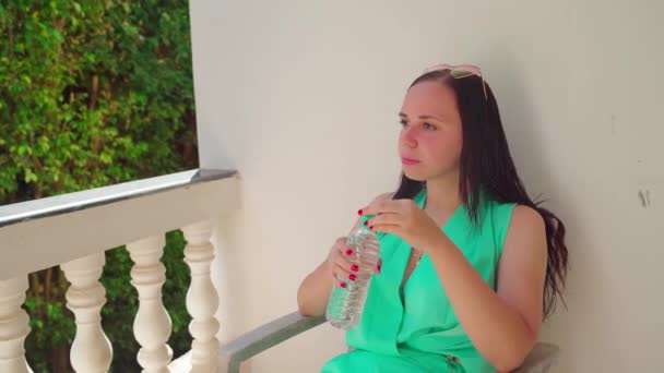 Una bella giovane donna è seduta su una sedia e beve un'acqua fredda da una bottiglia sulla terrazza. — Video Stock