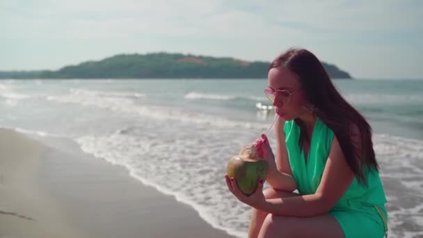 在阳光灿烂的日子里 一位美丽的年轻女子正坐在木头上 在海边或海边喝着椰子 — 图库视频影像
