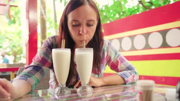 En vacker ung kvinna dricker hälsosamma smoothies i ett tropiskt café. Flickan dricker milkshakes med njutning. — Stockvideo