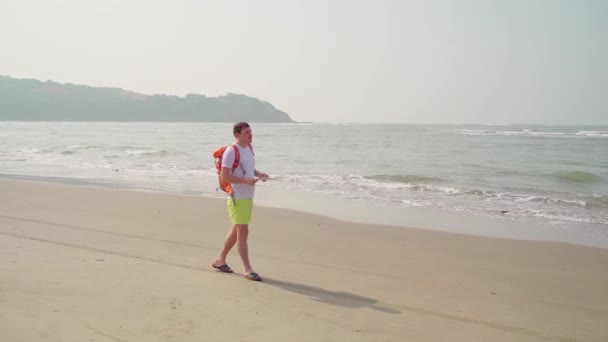 Traveling young man with backpack walking along sea coast. Male tourist reading and examining map while resting on sandy beach near waving sea. — Stock Video