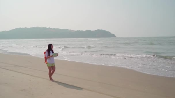 Eine reisende junge Frau mit Rucksack läuft an der Küste entlang. Touristin liest und untersucht Landkarte, während sie sich am Sandstrand in der Nähe des winkenden Meeres ausruht. — Stockvideo