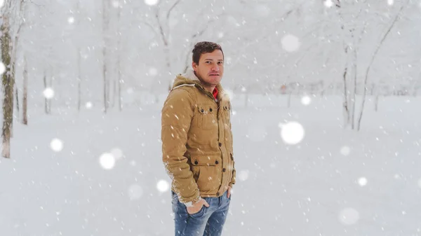 Retrato de jóvenes vestidos de chaqueta y vaqueros en temporada invernal. Un hombre agradable se pone de pie y escuche en el parque de invierno. La nieve fluida lo rodea todo.. —  Fotos de Stock