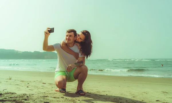 Casal feliz tomando selfies perto do mar. Casal amante abraçando durante a data na praia contra o mar acenando e céu sem nuvens. — Fotografia de Stock
