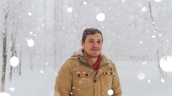 Retrato del joven en chaqueta durante la temporada invernal. Un hombre agradable se pone de pie y escuche en el parque de invierno. La nieve fluida lo rodea todo.. — Foto de Stock