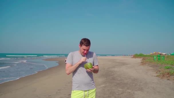 Розслаблений дорослий хлопець стоїть і п'є кокос на піщаному пляжі. Красивий чоловік насолоджується тропічним напоєм, просіюючи кокосову воду через солому біля моря . — стокове відео