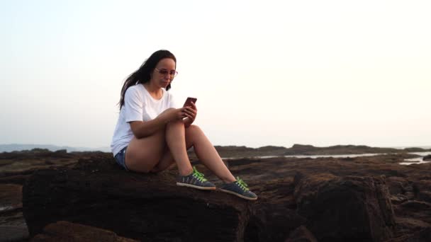 Mujer relajada sentada en piedra y hojeando las diversas noticias en el teléfono móvil en la orilla. Vista lateral de la agradable chica pasar tiempo disfrutando de vacaciones en la costa pedregosa . — Vídeos de Stock