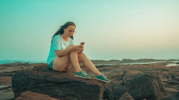 Relaxed woman sitting on stone and flipping through the various news in mobile phone on shore. Side view of pleasant girl spending time enjoying vacation on stony shore. — Stockfoto