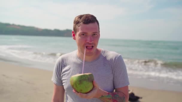 Relajado chico adulto de pie y beber coco en la playa de arena. Hombre guapo disfrutando de la bebida tropical, bebiendo agua de coco a través de paja cerca del mar . — Vídeos de Stock