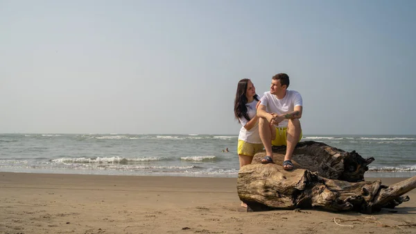 Casal feliz sentado no driftwood perto do mar. Amar casal abraçando e olhando uns para os outros enquanto sentado em log durante a data na praia contra o mar acenando e céu sem nuvens. — Fotografia de Stock