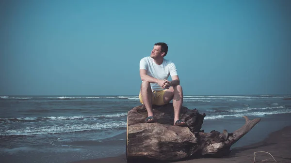 El hombre relajado se sienta en el tronco y mira hacia otro lado. Turista masculino se sienta en la madera a la deriva y entrecerran los ojos al sol brillante, descansando en la playa de arena cerca del mar ondulante . — Foto de Stock