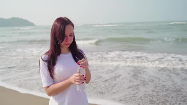 Giovane donna che beve acqua fredda dalla bottiglia vicino al mare. Adulti signora godendo di acqua minerale fresca sulla spiaggia sabbiosa. — Video Stock