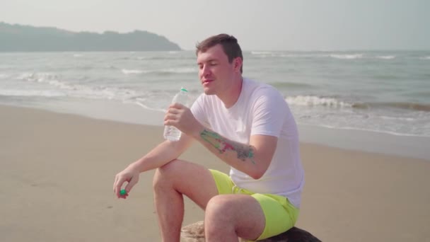 Jovem sentado no tronco e bebendo água fria de garrafa perto do mar. Adulto que gosta de água mineral fresca, sentado no driftwood na praia arenosa. — Vídeo de Stock