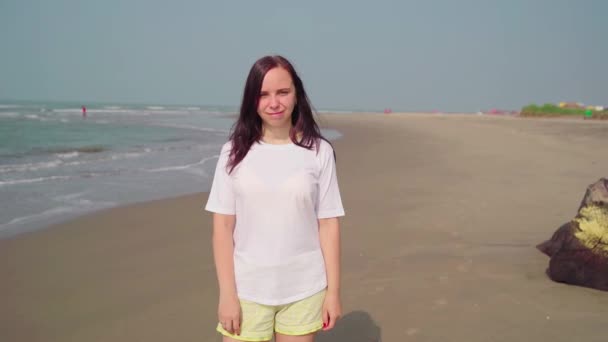 Όμορφη γυναίκα στέκεται στην αμμώδη παραλία κοντά στη θάλασσα. Γυναίκα τουρίστρια χαμογελά χαρούμενα στην ηλιόλουστη μέρα στην παραλία. — Αρχείο Βίντεο