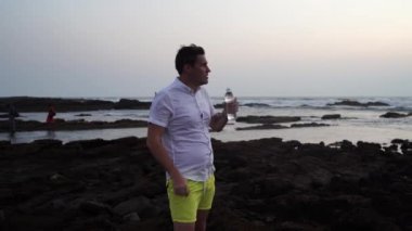 Kıyıda taş üstünde duran ve deniz kenarındaki şişeden soğuk su içen rahat bir adam. Yetişkin bir adamın serin maden suyunun tadını çıkarması, taşlı sahilde tatilin tadını çıkarması..