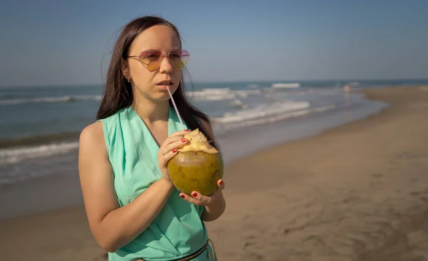 Mujer joven en gafas de sol bebiendo coco cerca del mar o el océano. Turista femenina disfrutando de bebidas tropicales, bebiendo agua de coco a través de paja en la playa de arena . — Foto de Stock