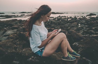 Relaxed woman sitting on stone and flipping through the various news in mobile phone on shore. Side view of pleasant girl spending time enjoying vacation on stony shore.