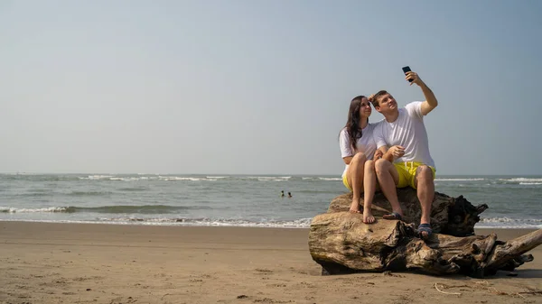 Glückliches Paar, das auf Treibholz am Meer sitzt. Liebespaar umarmt sich und macht Selfies beim Date am Strand vor winkendem Meer und wolkenlosem Himmel. — Stockfoto