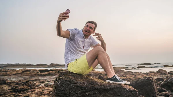 Homem relaxado sentado em pedra e tirando selfie no smartphone em terra. Vista lateral do homem agradável passar o tempo desfrutando de férias e fotografar de si mesmo no telefone celular na costa pedregosa . — Fotografia de Stock