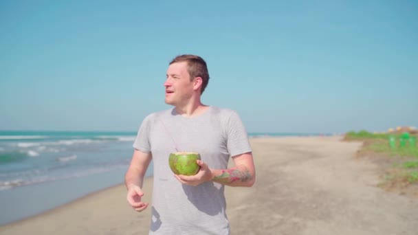 Relaxed adult guy standing and drinking coconut on sandy beach. Handsome man enjoying tropical drink, sipping coconut water through straw near sea. — Wideo stockowe