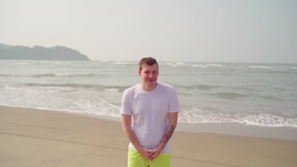 El hombre hermoso se encuentra en la playa de arena cerca del mar. El turismo masculino sonríe alegremente en un día soleado en la costa.. — Vídeos de Stock