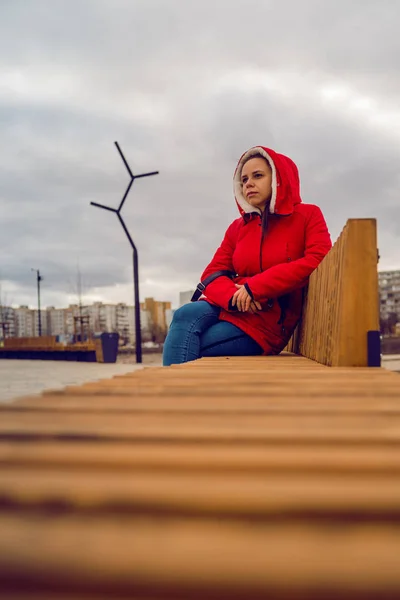 Young woman in red jacket and hood sitting on wooden bench in cloudy weather. Adult lady looking thoughtfully into distance. — Φωτογραφία Αρχείου