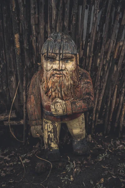 Gnome de madeira perto de cerca de madeira no chão. Velha figura rachada esculpida em madeira. Jardim gnomo no campo . — Fotografia de Stock