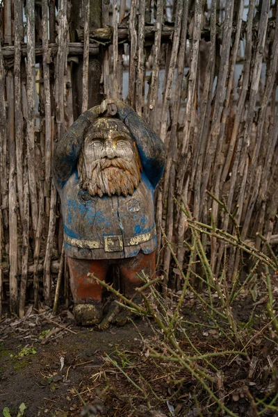 Gnome de madeira perto de cerca de madeira no chão. Velha figura rachada esculpida em madeira. Jardim gnomo no campo . — Fotografia de Stock