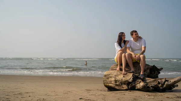 Happy couple posing on driftwood near sea. Loving couple hugging during date on beach against waving sea and cloudless sky. — Stock Photo, Image