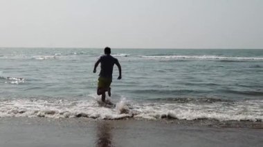 Deniz kıyısında bir erkek. Denizde su sıçratan ve kameraya poz veren çekici bir adam. Gülümseyen adam deniz suyunda eğleniyor..