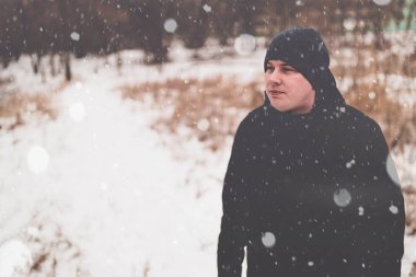 Adult full man standing in park in winter season. Serious young male wearing black jacket and hat, standing and looking away in snowy day.