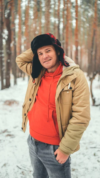 Knappe man die in de winter in het bos staat. Brutale jonge man draagt beige jas over rode hoodie en oorflap hoed poseren in besneeuwd bos. — Stockfoto