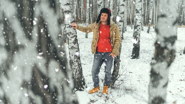 Hombre guapo de pie en el bosque en temporada de invierno. Brutal joven macho vistiendo chaqueta beige sobre sudadera con capucha roja y sombrero de oreja posando en el bosque nevado . —  Fotos de Stock