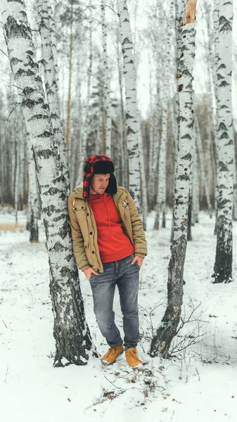 Handsome man standing in forest in winter season. Brutal young male wearing beige jacket over red hoodie and earflap hat posing in snowy forest. — Stock Photo, Image