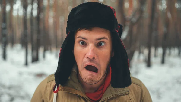 Az ember grimaszol az erdőben a téli szezonban. Portré őrült férfi fülbevaló kalap és grimaszoló arc közelről a téli erdőben. — Stock Fotó