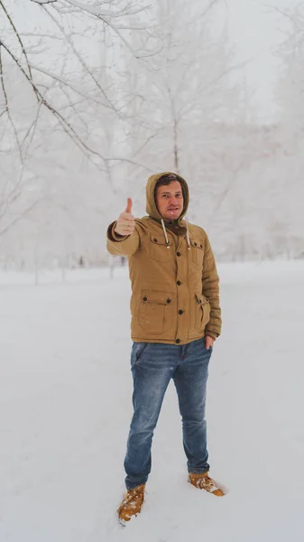冬天,一个英俊的男人站在公园里,竖起大拇指.在雪天，身穿米色帽衫的成年男青年表现出认可的姿态. — 图库照片