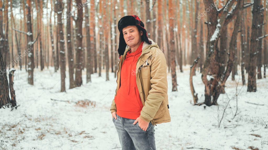 Handsome man standing in forest in winter season. Brutal young male wearing beige jacket over red hoodie and earflap hat posing in snowy forest.