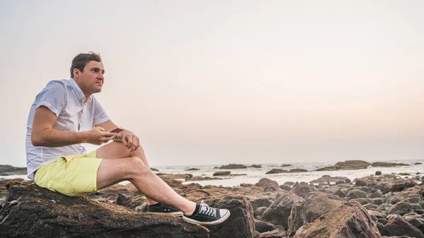 Hombre relajado sentado en piedra y hojeando las diversas noticias en el teléfono móvil en la orilla. Vista lateral de tipo agradable pasar tiempo disfrutando de vacaciones en la costa pedregosa . — Foto de Stock