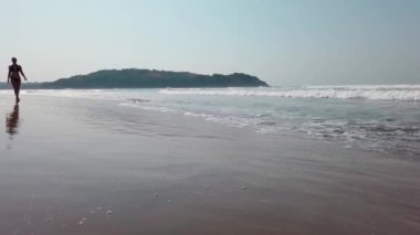 Relaxed woman walking along sea coast in sunny day. Female tourist spending time enjoying vacation and promenading on sandy beach near waving sea.