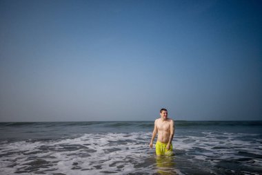 Deniz kıyısında bir erkek. Denizde su sıçratan ve kameraya poz veren çekici bir adam. Gülümseyen adam deniz suyunda eğleniyor..
