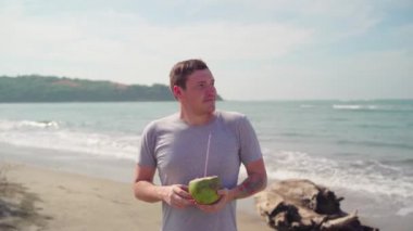 Relaxed adult guy standing and drinking coconut on sandy beach. Handsome man enjoying tropical drink, sipping coconut water through straw near sea.