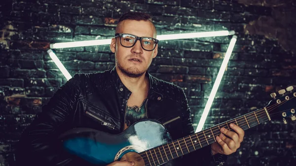 Stylish guy with bird tattoo on chest looking away and playing guitar while standing against shabby brick wall with triangle illumination