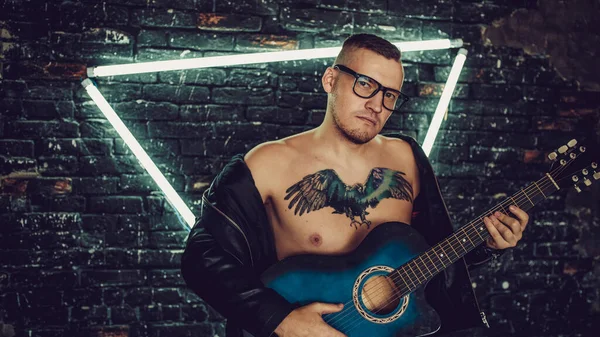 Tattooed man playing guitar near illuminated wall. Stylish guy with bird tattoo on chest looking away and playing guitar while standing against shabby brick wall with triangle illumination